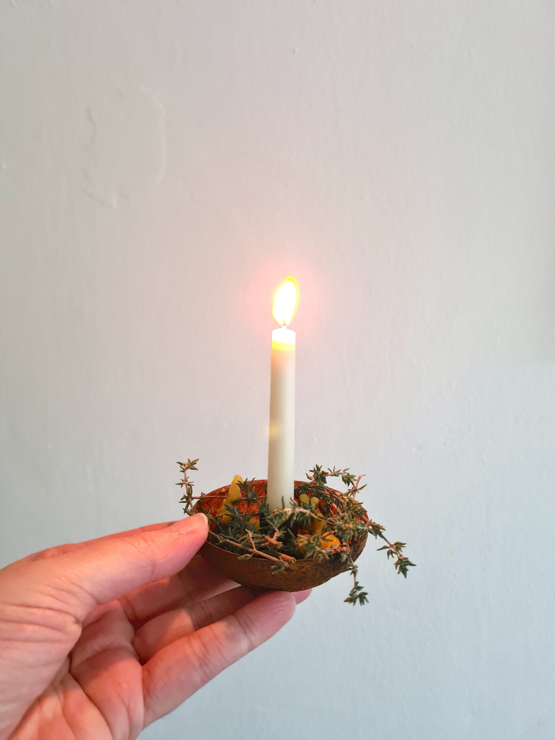 Een hand die een klein, rond en bruin kandelaartje vasthoudt. Op de bodem van de kandelaar liggen kleine droogbloemetjes, druppels gele bijenwas en takjes tijm. Verticaal in de kandelaar staat een klein wit kaarsje. Hij staat aan, het vlammetje schijnt. Alles op een witte muur als achtergrond, gemaakt door Natuurlijk Verwonderen.